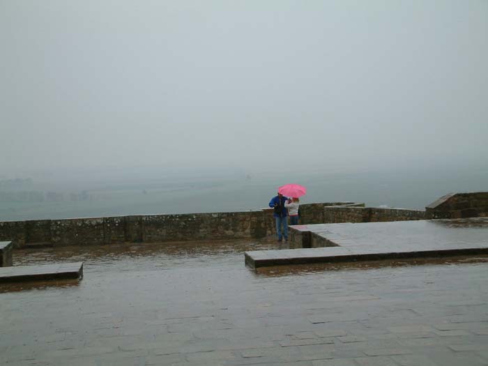 A couple is standing& on the large terrace in front of the abbey church. She wears a pink umbrella to protect herself from the inconvenience of the bad weather.