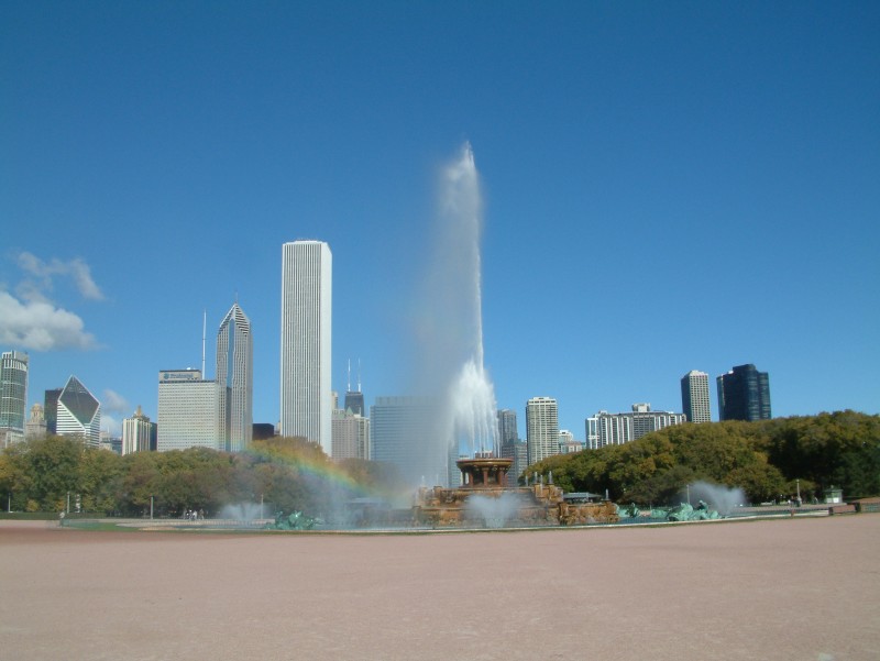 Clarence Buckingham Brunnen mit einem kleinen Regenbogen. Im Hintergrund ist die beeindruckende Skyline der Innenstadt von Chicago zu sehen. Die Hochhäuser überragen das Grün des Grant Parks.