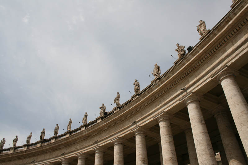 Der eliptische Petersplatz ist auf& zwei Seiten von Säulengängen eingeschlossen. Architekt Bernini selbst sagte, dass "die Kirche Petri sozusagen die Mutter aller anderen Kirchen ist und sie daher Kolonnaden haben muss, die wie mit mütterlich ausgebreiteten Armen die Katholiken aufnehmen, um sie in ihrem Glauben zu bestärken, die Irrgläubigen, um sie der Kirche zuzuführen und die Ungläubigen, um sie im wahren Glauben zu erleuchten".